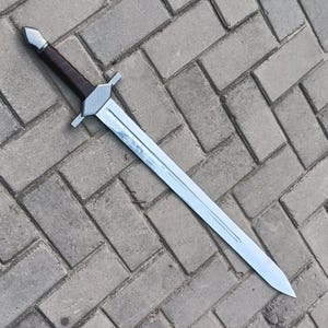 May include: A silver and brown sword with a straight blade and a crossguard. The handle is wrapped in brown material. The sword is lying on a gray brick surface. The sword appears to be a replica or prop.