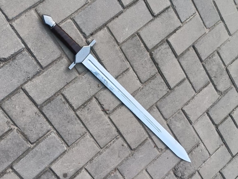 May include: A silver and brown sword with a straight blade and a crossguard. The handle is wrapped in brown material. The sword is lying on a gray brick surface. The sword appears to be a replica or prop.
