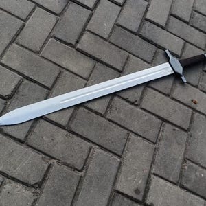 May include: A long, silver sword with a dark brown handle and a crossguard. The blade is straight and sharp, reflecting the light. The sword is lying on a gray brick surface.
