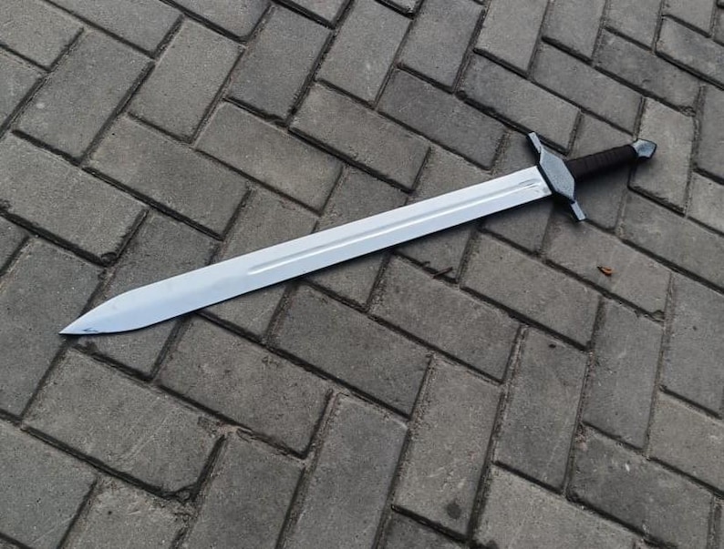 May include: A long, silver sword with a dark brown handle and a crossguard. The blade is straight and sharp, reflecting the light. The sword is lying on a gray brick surface.