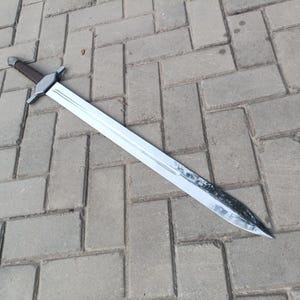 May include: A long, silver sword with a brown handle and a crossguard. The blade is shiny and appears to be made of metal. The sword is lying on a gray brick surface.