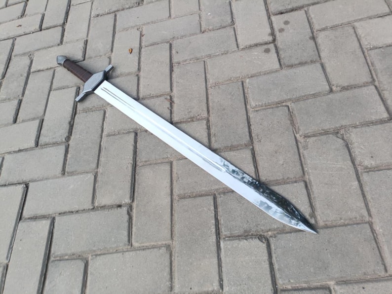 May include: A long, silver sword with a brown handle and a crossguard. The blade is shiny and appears to be made of metal. The sword is lying on a gray brick surface.
