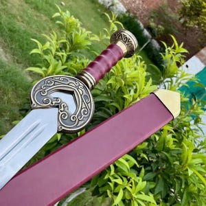 May include: A decorative sword with a silver blade and a burgundy handle. The handle is wrapped in a textured material. The guard is a circular design with ornate gold detailing. The scabbard is burgundy with a gold tip.