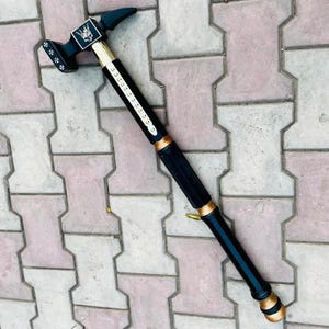 Può includere: Un martello nero e oro con un design decorativo sulla testa. Il martello ha un lungo manico nero con dettagli dorati e un emblema decorativo. La testa del martello ha un artiglio e una superficie di battuta.