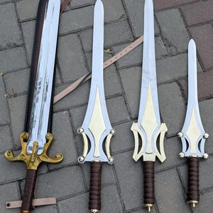 May include: Collection of four swords with silver blades and gold-colored hilts. The hilts have intricate designs and brown leather-wrapped handles. One sword is in a black sheath. The swords vary in size, with the largest measuring approximately 36 inches.