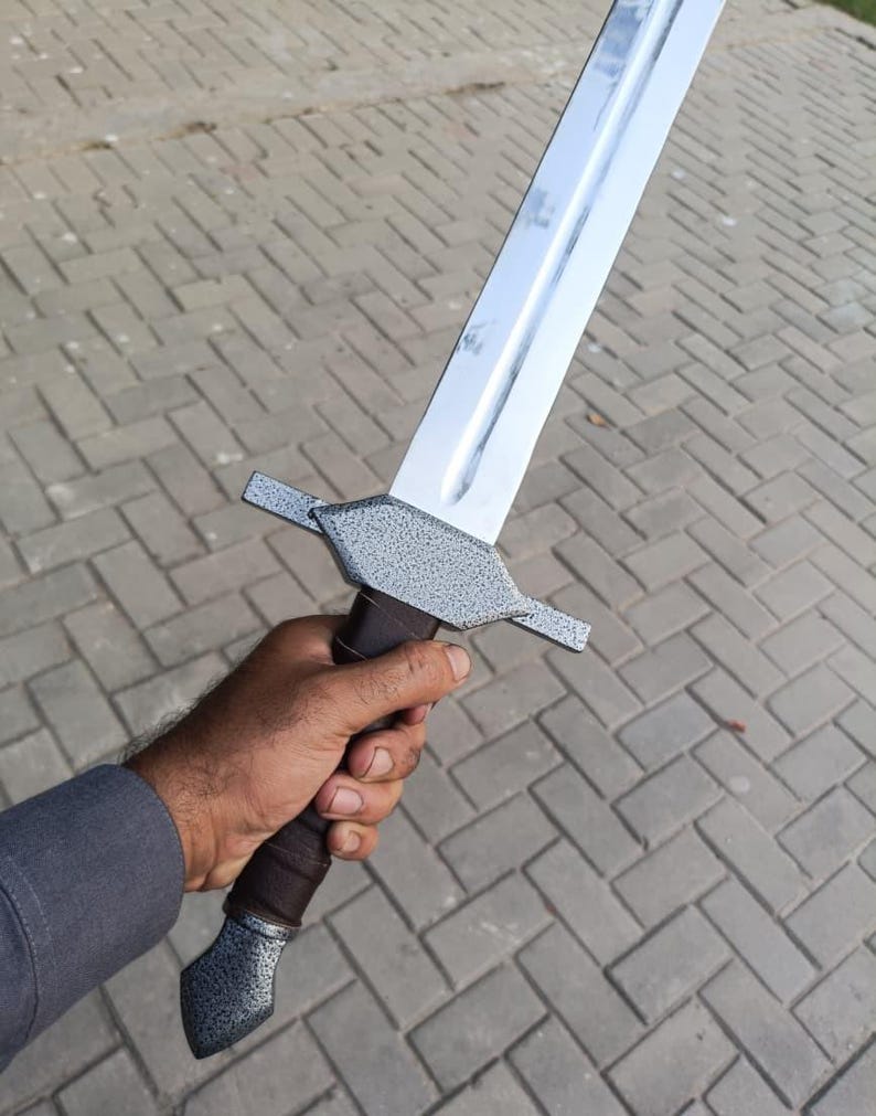 May include: A silver and gray sword with a brown handle. The blade is long and shiny, and the handle has a textured grip. The sword has a crossguard and a pommel. The sword is held in a hand.