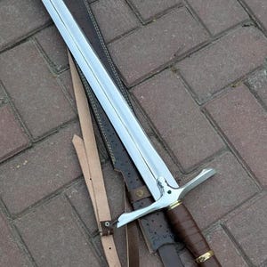 Op de afbeelding: Een lang, zilveren zwaard met een bruin, met leer omwikkeld handvat en een bijpassende schede. Het zwaard heeft een kruisvormige pareerstang en een leren riem. De schede is donkerbruin met decoratieve stiksels. Het zwaard ligt op een bakstenen oppervlak.