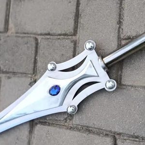 May include: A silver-colored sword with a decorative hilt featuring a blue gem and rounded studs. The blade is long and pointed, and the sword rests on a gray brick surface. The sword's design is ornate.