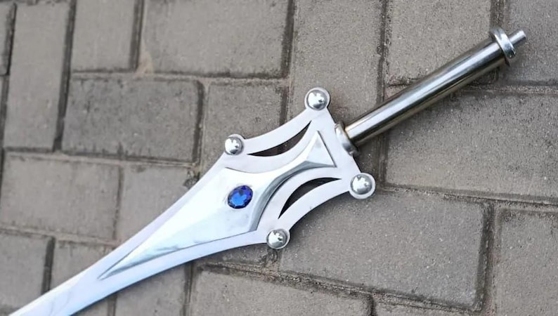May include: A silver-colored sword with a decorative hilt featuring a blue gem and rounded studs. The blade is long and pointed, and the sword rests on a gray brick surface. The sword's design is ornate.