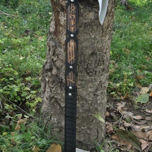 May include: A Viking-style axe with a polished steel blade and a wooden handle. The handle is wrapped in black leather and adorned with silver rivets and etched runes. The axe is displayed outdoors against a backdrop of greenery.