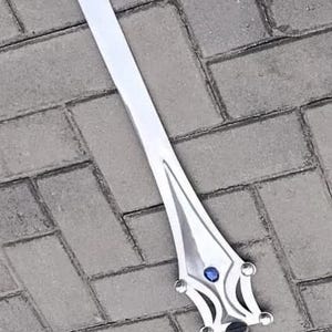 May include: A silver-colored sword with a long, pointed blade. The hilt features a decorative design with a blue gem in the center. The sword is lying on a gray brick surface.