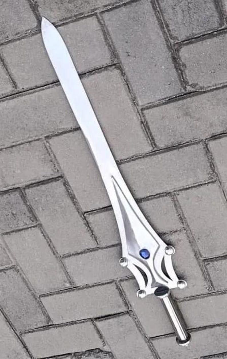 May include: A silver-colored sword with a long, pointed blade. The hilt features a decorative design with a blue gem in the center. The sword is lying on a gray brick surface.
