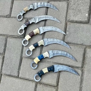 May include: Six curved karambit knives with patterned steel blades and various handle colors, including cream, brown, and blue. Each knife features a finger ring at the base of the handle. The knives are arranged on a gray brick surface.