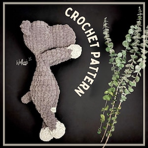 Puede incluir: Un juguete de hipopótamo de crochet gris con pies blancos. El juguete está acostado sobre una superficie negra con una rama de hojas de eucalipto en el fondo. El texto "CROCHET PATTERN" está escrito en blanco sobre fondo negro.