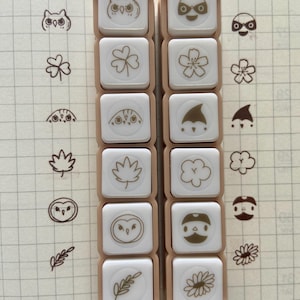 Peut inclure: Deux rangées de tampons carrés avec divers motifs sur le thème de la nature, notamment des hiboux, des trèfles, des fleurs et des feuilles. Les tampons ont une surface carrée blanche avec un bord beige. Les motifs sont à l'encre brune. Le fond est un papier quadrillé.