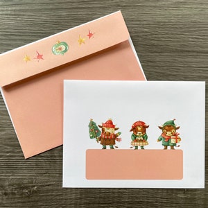 Peut inclure: Une carte blanche avec une enveloppe rose. La carte représente trois vaches de dessin animé portant des chapeaux d'hiver et tenant des cadeaux. La carte a une bannière rose en bas avec de l'espace pour un message.