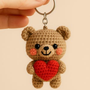 May include: A crocheted brown bear keychain holding a red heart. The bear has black eyes and pink cheeks. The text "Valentine Bear Keychain Crochet Pattern" is displayed below the bear.