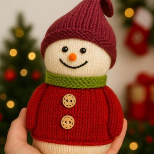May include: A knitted snowman figure with a burgundy hat, green scarf, and red sweater. The snowman has a cream-coloured body, black eyes, an orange nose, and a stitched smile. Two wooden buttons fasten the sweater.