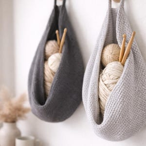 May include: Two hanging knitted storage bags, one gray and one light gray, each filled with yarn balls and wooden knitting needles. The bags are suspended from wooden pegs on a white wall, creating a cozy, organized craft space.