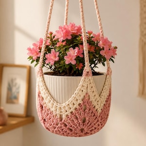 May include: A hanging plant holder made of crocheted yarn in shades of cream and dusty rose, featuring a V-shaped pattern. The holder contains a white, vertically ribbed ceramic pot filled with a blooming azalea plant with bright pink flowers.