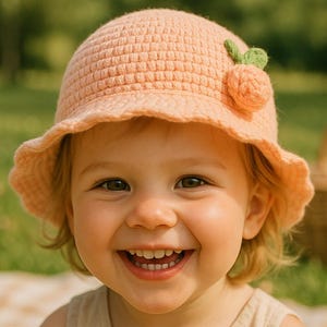 Op de afbeelding: Een lachend kind draagt een perzikroze gehaakte hoed met een groene en perzikroze fruitversiering. De hoed heeft een geschulpte rand. Het kind draagt een beige tanktop. Een picknickmand staat op de achtergrond.