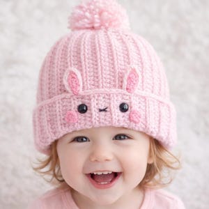 May include: A light pink knitted beanie hat with a bunny face design. The hat has a pom-pom on top, with embroidered bunny ears, eyes, and a nose. The beanie is designed for babies and toddlers.