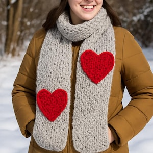 May include: A grey knitted scarf featuring two large red heart appliqués. The scarf is worn with a navy blue knitted hat and a brown jacket. The background shows a snowy, outdoor scene.