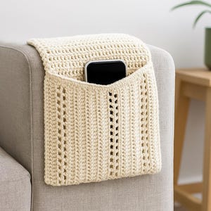 Könnte beinhalten: Eine cremefarbene, gehäkelte Sofarmtasche mit einem schwarzen Smartphone darin. Die Tasche ist am Arm eines grauen Sofas befestigt. Das Häkeldesign weist ein strukturiertes Muster mit durchbrochenen Details auf.