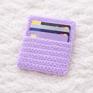 May include: A handmade, lavender-colored crochet card holder. The small, rectangular wallet is filled with several credit cards. The crochet stitch creates a textured surface. The card holder is displayed on a white, fluffy background.