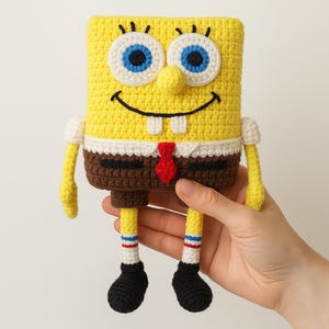 May include: A yellow, brown, and white crocheted plush toy shaped like a rectangular character with large white eyes, blue irises, a black smile, two white teeth, a red tie, and black shoes. The figure has thin yellow arms and legs.