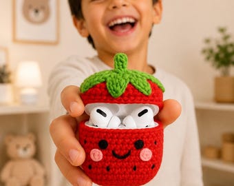 May include: A red strawberry-shaped crochet case for wireless earbuds. The case features a green stem top, a smiling face with black eyes, a curved mouth, and pink cheeks. The case is open, revealing white wireless earbuds inside.