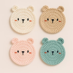 Può includere: Quattro sottobicchieri all'uncinetto fatti a mano a forma di faccia di orso. I sottobicchieri sono color crema, beige, rosa e verde acqua. Ognuno ha un naso nero, piccoli occhi neri e guance rosse.
