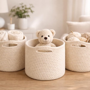 Könnte beinhalten: Drei cremefarbene, gehäkelte Aufbewahrungskörbe mit Griffen. Ein Korb enthält gerollte Handtücher, ein anderer einen Teddybären und der dritte Wolle und Stricknadeln. Die Körbe stehen auf einer Holzoberfläche.