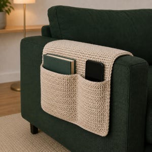 May include: A beige crochet sofa armrest organiser featuring two deep pockets draped over the side of a dark green couch. The left pocket contains a dark green hardback book, while the right pocket holds a black smartphone, providing storage.