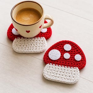 May include: A cup of coffee rests on a red and white mushroom-shaped crochet coaster. The coaster has a red top with white circles and a white, rectangular base. The cup is beige with red heart accents.