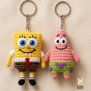 Könnte beinhalten: Zwei Häkel-Schlüsselanhänger, die SpongeBob Schwammkopf und Patrick Star darstellen. Die SpongeBob-Figur ist gelb, braun und weiß mit blauen Augen. Die Patrick-Figur ist rosa und hellgrün mit violetten Flecken. Beide sind an silbernen Metallringen befestigt.