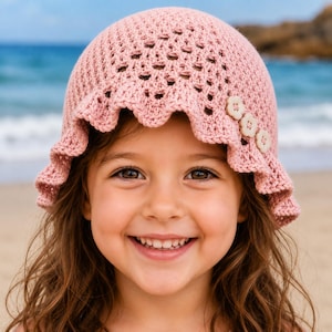 May include: A pale pink crocheted sun hat with a frilled edge and three decorative buttons. The hat is worn by a young child with brown eyes and long brown hair. The background shows a beach scene with sand and water.