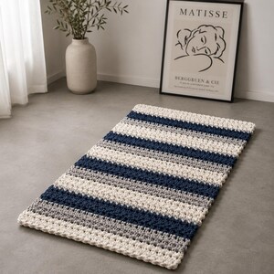 Può includere: Un tappeto rettangolare all'uncinetto caratterizzato da un motivo a righe orizzontali ripetute nei colori alternati crema, grigio chiaro e blu navy scuro. Il tappeto ha una struttura in filato spesso e testurizzato con un punto uniforme.