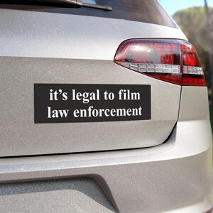 May include: A black rectangular car magnet with the white text "it's legal to film law enforcement" is affixed to the rear of a silver car. The car's taillight is visible on the right side of the image.