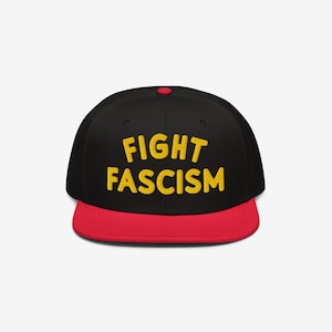 May include: Black baseball cap with a red brim. The cap features the yellow text "FIGHT FASCISM" across the front. The cap has a red button on top.