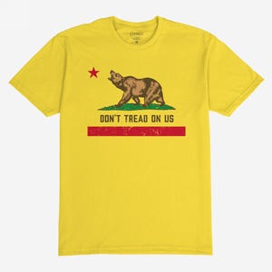 May include: Yellow t-shirt featuring a brown bear, red star, and the text "DON'T TREAD ON US." The bear is standing on green grass, with a red stripe below the text. The shirt is a classic crew neck.
