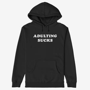May include: Black hooded sweatshirt with the text "ADULTING SUCKS" in white, bold, uppercase letters. The sweatshirt has a front pocket and a drawstring hood. The garment is made of a soft material.