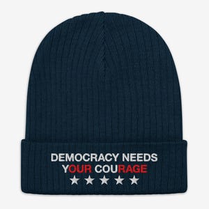Puede incluir: Un gorro de punto acanalado azul marino con el texto bordado "DEMOCRACY NEEDS YOUR COURAGE" en blanco y rojo, con cinco estrellas blancas debajo. Un accesorio de invierno.