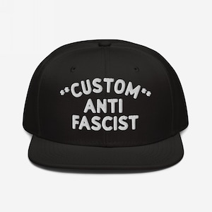 May include: Black flat-brimmed baseball cap with the white text "CUSTOM ANTI FASCIST" embroidered across the front. The text is in a bold, sans-serif font, with asterisks on either side of the word "CUSTOM".