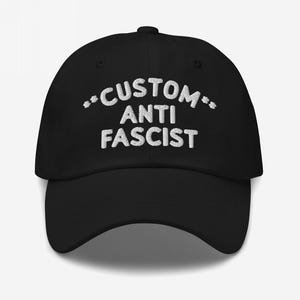 Front view of black unstructured dad hat with “Custom Anti Fascist” embroidered in white thread.