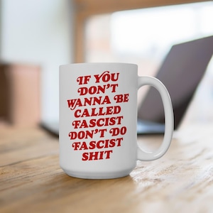 May include: White ceramic mug with a large handle. The mug features red text that reads: "IF YOU DON'T WANNA BE CALLED FASCIST DON'T DO FASCIST SHIT". The mug is on a wooden surface with a laptop in the background.