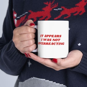 May include: A white ceramic mug with the text "IT APPEARS I WAS NOT OVERREACTING" in red. The mug is held by a person wearing a dark blue sweater with a red reindeer pattern.