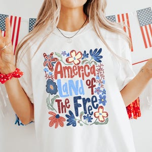 May include: White t-shirt with the text "America Land of The Free" in red, blue, and green floral design. The image is decorated with American flags and red, white, and blue decorations. A person is holding a drink with a strawberry.