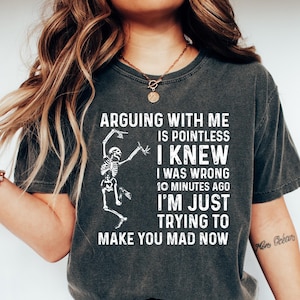 May include: Dark gray t-shirt with the text "ARGUING WITH ME IS POINTLESS I KNEW I WAS WRONG 10 MINUTES AGO I'M JUST TRYING TO MAKE YOU MAD NOW" and a skeleton graphic. The shirt is paired with blue jeans and white shoes.