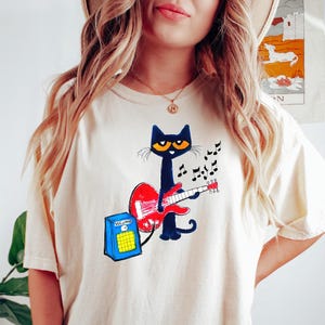 May include: Cream-colored t-shirt featuring a cartoon black cat playing a red guitar. The cat has yellow eyes and is surrounded by musical notes. A blue amplifier with the word "VOLUME" is next to the cat. The shirt is a casual, relaxed fit.